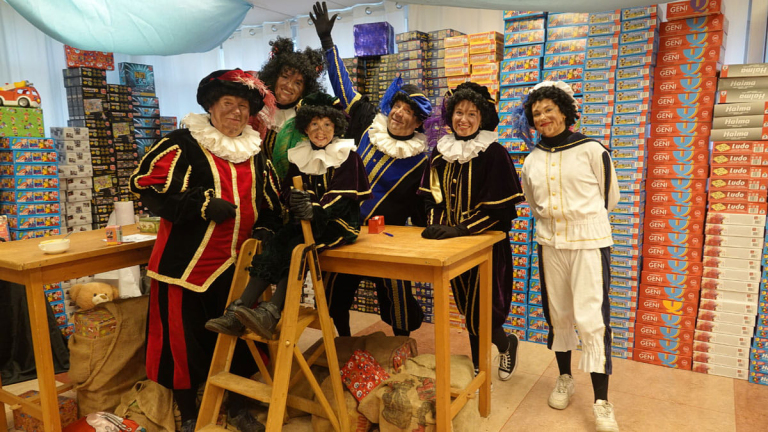 Groep mensen gekleed in traditionele kostuums met kleurrijke outfits en zwarte schmink op hun gezichten, staande naast tafels in een ruimte gevuld met stapels speldozen.