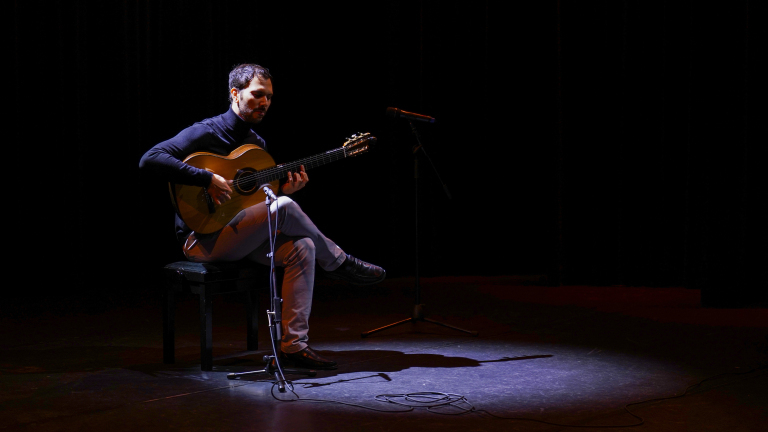 Een man zit op een stoel op een podium en speelt gitaar onder een spotlicht, met een microfoon voor hem.