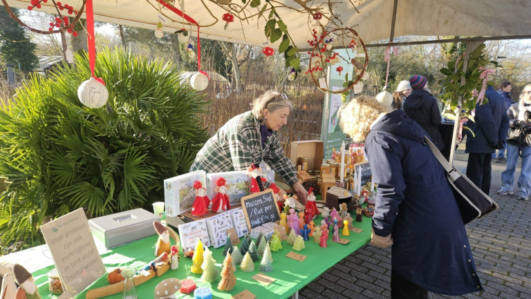 Twee mensen staan bij een kraampje op een markt met kerstdecoraties en handgemaakte kaarsen uitgestald op een groene tafel, omgeven door planten en bezoekers.
