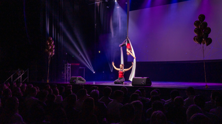 Twee acrobaten voeren op een podium een luchtacrobatiekact uit met witte doeken, terwijl een publiek toekijkt; de scène is kleurrijk verlicht met spotlights en er hangen ballonnen aan beide zijkanten van het podium.