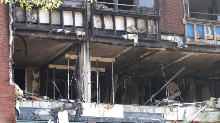 Close-up van de schade aan een gebouw na een brand, met zwartgeblakerde muren en gesmolten isolatiemateriaal, gescheurde ramen en blootgelegde interne structuren.