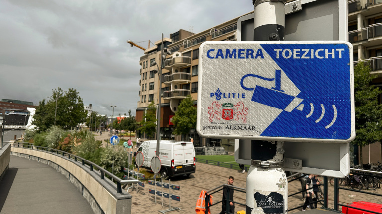 Bord met de tekst 'Camera Toezicht' in Alkmaar, met politie en gemeente logo's, tegen een stedelijke achtergrond met een gebouw en een busje.