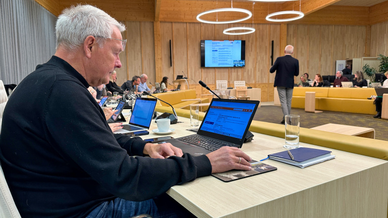 Een man met grijs haar zit aan een tafel en werkt op zijn laptop, terwijl hij met anderen in een moderne vergaderruimte zit. Er hangen cirkelvormige lampen aan het plafond en in de achtergrond spreekt een persoon voor een beeldscherm.