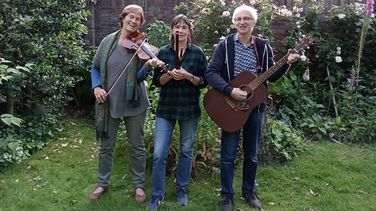 Drie personen staan in een tuin met muziekinstrumenten; de linker persoon speelt viool, de middelste houdt een houten fluit en de rechter persoon heeft een gitaar.