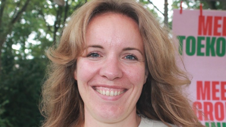 Vrouw met lang lichtbruin haar glimlacht naar de camera, met een groene achtergrond en een bord met tekst op de achtergrond.