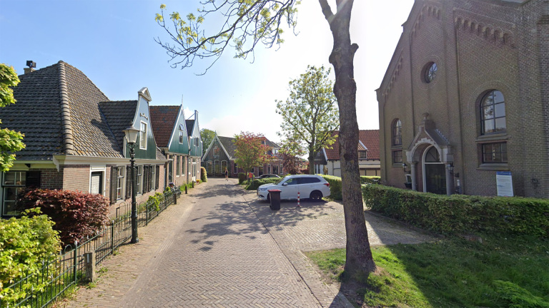 Een straat in een Nederlands dorp met traditioneel vormgegeven huizen met puntgevels aan de linkerzijde en een groot bakstenen gebouw aan de rechterzijde. Er is een boom, een geparkeerde auto en een met struiken omheind gebied zichtbaar.