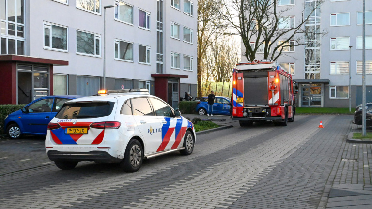 Politie- en brandweerwagen staan geparkeerd voor een wooncomplex met meerdere verdiepingen. De voertuigen hebben waarschuwingslichten aan. Er zijn een paar mensen in de buurt van de gebouwen aanwezig.