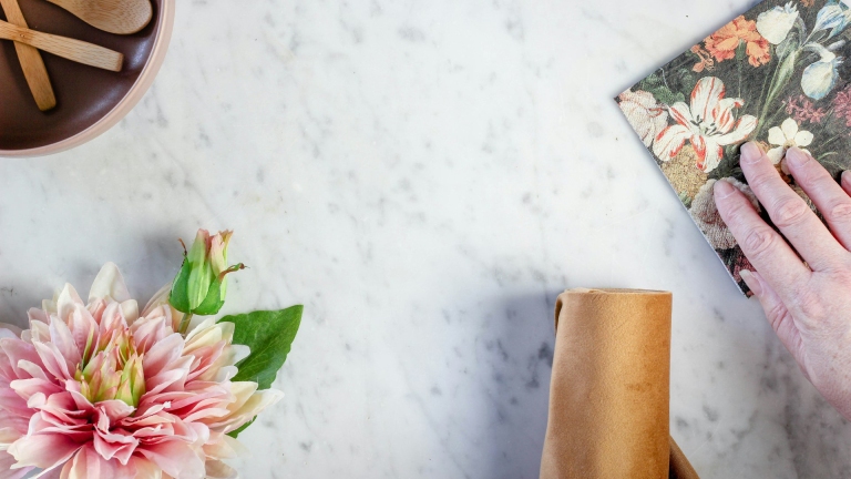 Bovenaanzicht van een marmeren tafel met een hand die een boek met bloemenachtergrond vasthoudt, een roze bloem met groene bladeren links, een bruine stoffen servet rechtsonder en een bruine kom met houten lepels linksboven.