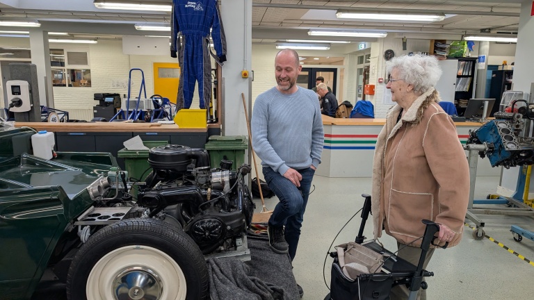 Twee mensen in een werkplaats staan bij een oldtimer auto met een open motorkap, omgeven door gereedschap en werkbanken.