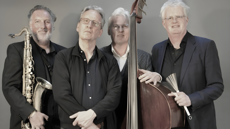 Vier mannen poseren samen voor een neutrale achtergrond met muzikale instrumenten, waaronder een saxofoon en een contrabas.