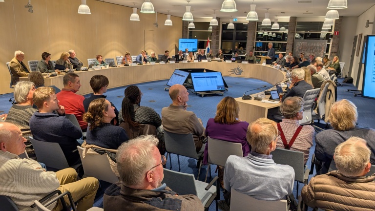 Een gemeenteraadsvergadering in een moderne zaal met deelnemers die rond een ovale tafel zitten, omringd door publiek dat op stoelen zit. Grootschermen tonen informatie, en de zaal heeft een professionele uitstraling met veel verlichting.