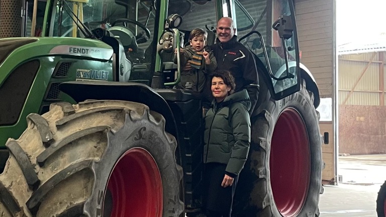 Een man, een vrouw en een kind staan naast een grote groene tractor in een schuur, met de man die het kind vasthoudt.