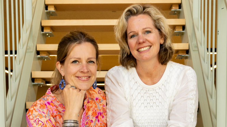 Twee vrouwen zitten glimlachend naast elkaar op een houten trap.