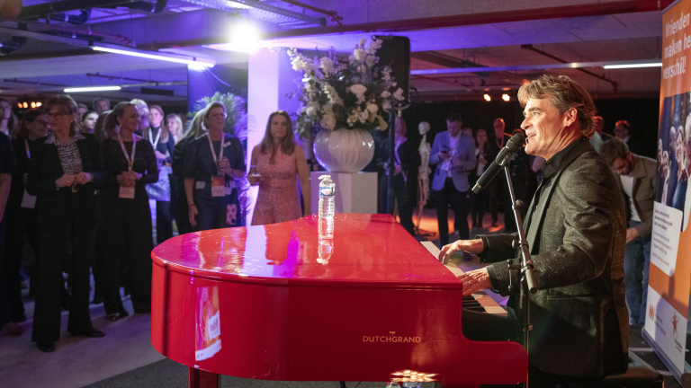 Man speelt piano en zingt voor een staand publiek tijdens een evenement; de piano is felrood gekleurd.