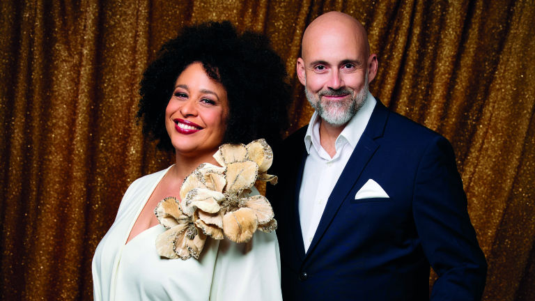 Twee mensen poseren voor een glinsterende gouden achtergrond; de vrouw draagt een witte jurk met een grote bloemenversiering en de man een donker pak met een wit overhemd en pochet.