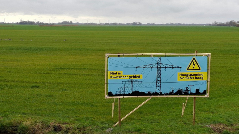 Waarschuwingsbord in een groene weide met tekst "Niet in kwetsbaar gebied. Hoogspanning 62 meter hoog" en een afbeelding van hoogspanningsmasten.
