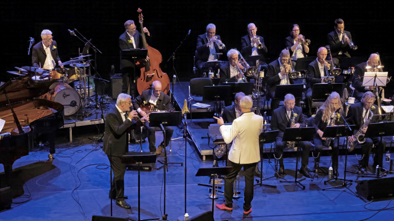 Een bigband speelt op een podium, begeleid door een dirigent in een witte jasje. De muzikanten bespelen verschillende instrumenten, waaronder trompetten, saxofoons, een contrabas, en een drumstel. Het podium is rijk gevuld met bladmuziekstandaarden en andere muziekinstrumenten.