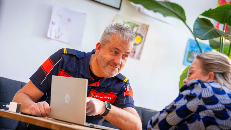 Brandweerman in uniform zit met vrouw aan tafel, kijkend naar een laptop in een woonkameromgeving met planten en muurdecoraties.