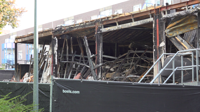 Uitgebrand gebouw met ingestorte structuur en zwartgeblakerde resten, omliggende gebouwen op de achtergrond.