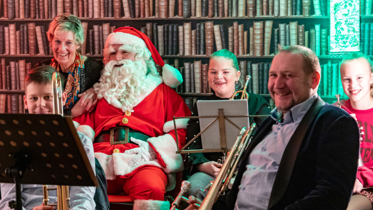 Groep mensen poseert met een persoon verkleed als de Kerstman, zittend in een ruimte met een boekenwand op de achtergrond.