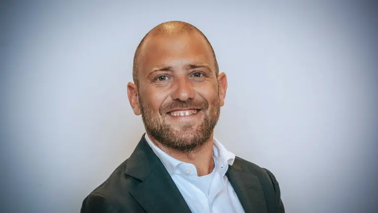 Man met een kale kop en baard, gekleed in een donker pak en witte overhemd, glimlacht tegen een lichte achtergrond.