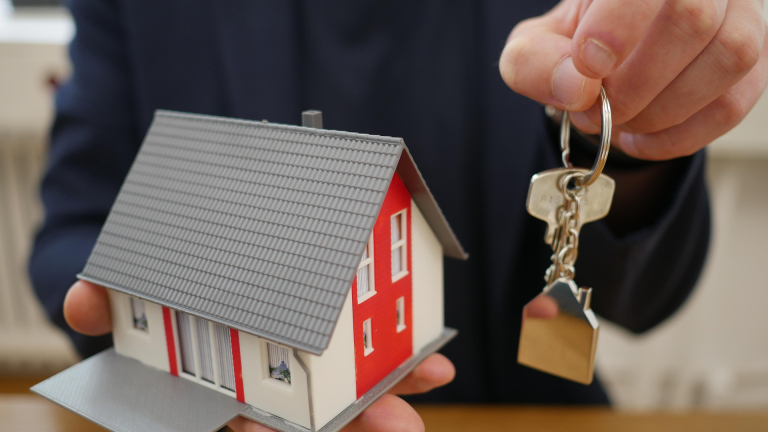Een hand houdt een miniatuurhuis en een sleutelbos met huissleutels vast, symboliserend een woningoverdracht.