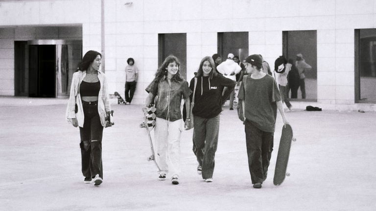 Vier jongeren lopen met skateboards in een open ruimte, lachend en pratend, met een gebouw op de achtergrond.