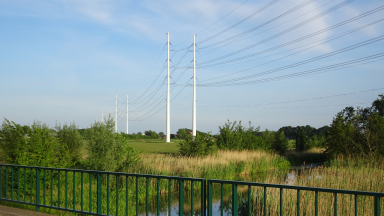 Elektriciteitsmasten in een groen landschap met bomen en een kleine waterweg，vooraan een groene reling.