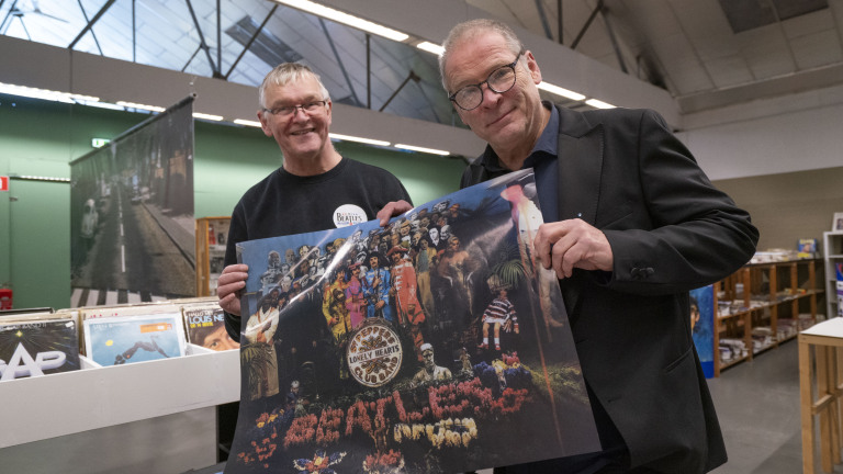 Twee mannen houden een poster vast van het album "Sgt. Pepper's Lonely Hearts Club Band" in een platenwinkel.