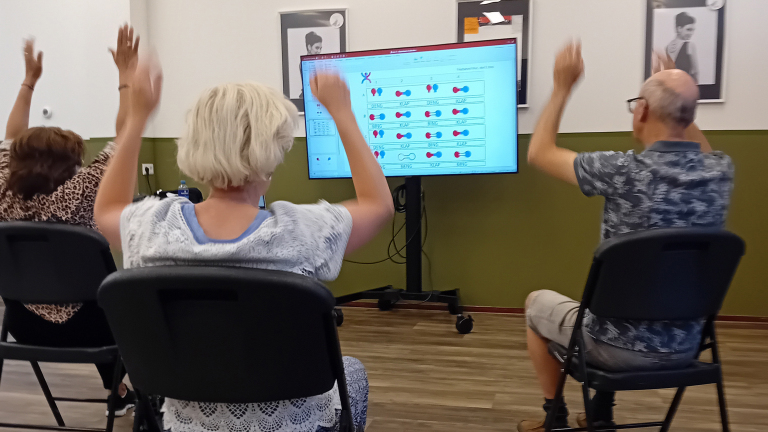 Drie personen zitten op stoelen in een ruimte en hebben hun armen opgeheven, gericht op een groot scherm met een interactieve presentatie op de achtergrond.