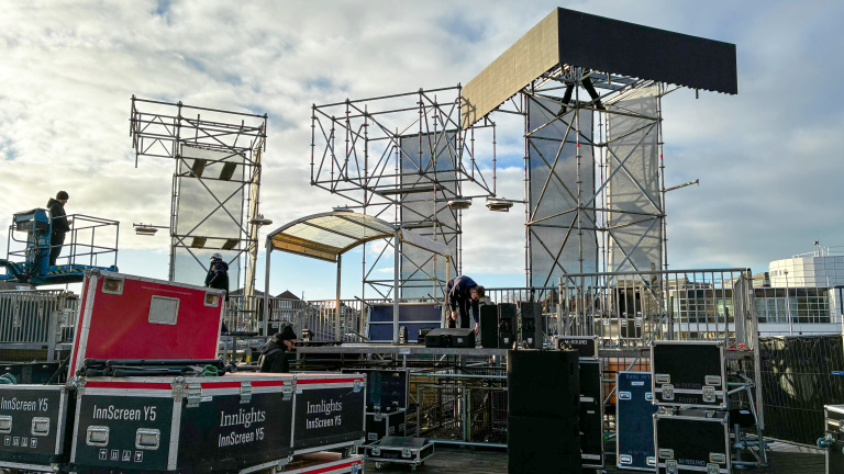 Mensen bouwen een groot podium met metalen steigers en schermen, omringd door apparatuur en apparatuurkisten.
