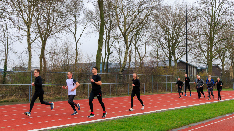 Een groep mensen rent op een atletiekbaan, omgeven door kale bomen op een grijze dag.