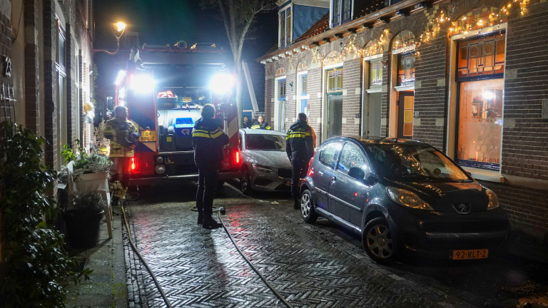 Brandweerlieden en politie bij een brandweerwagen in een smalle straat, met een geparkeerde auto ernaast en feestverlichting aan het huis.