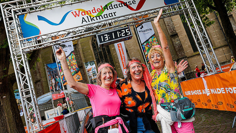 Drie lachende vrouwen poseren met opgeheven handen bij de finish van de Wandel4daagse Alkmaar, onder een spandoek met een klok die 13:11:25 aangeeft.