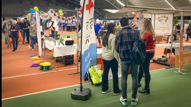 Mensen bezoeken stands bij een indoor evenement, met zichtbare Rode Kruis en Leergeld banners.