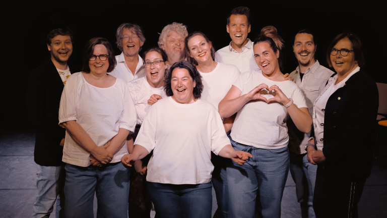 Een groep van elf personen poseert lachend voor de camera, sommigen dragen witte bovenkleding, een persoon maakt een hartvorm met de handen.