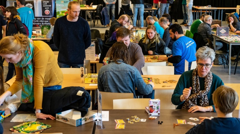 Mensen spelen bordspellen aan tafels tijdens een evenement in een zaal met verschillende stands.