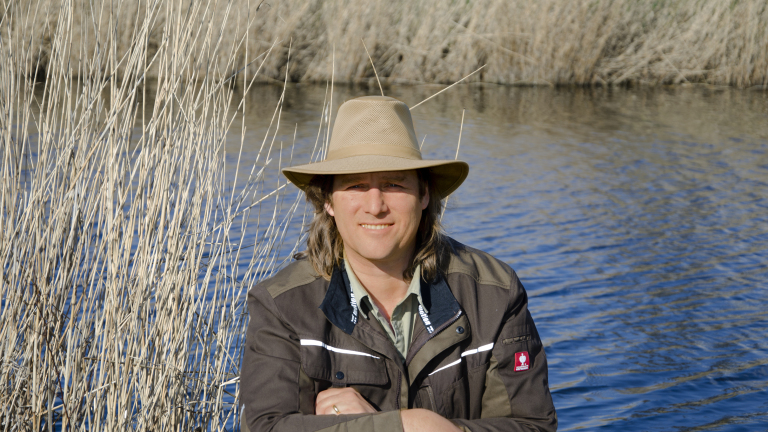 Persoon met hoed en jas bij riet en een watergebied, glimlachend naar de camera.
