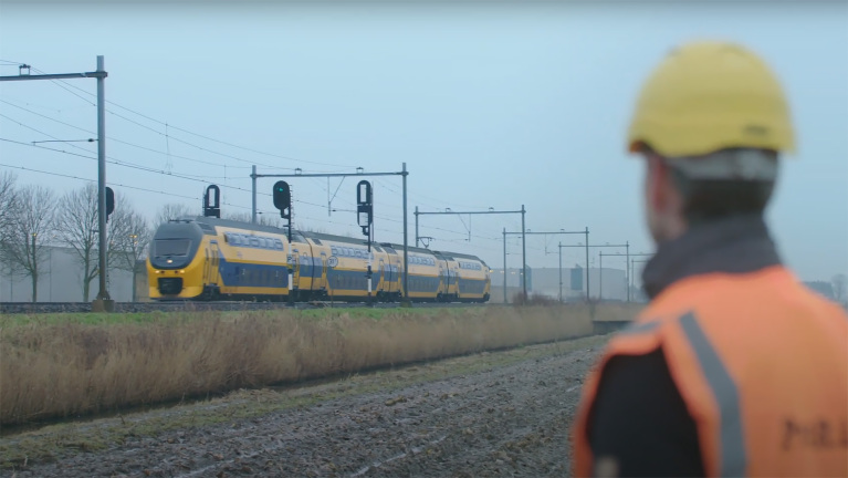 Een man met veiligheidshelm kijkt naar een passerende gele trein op het spoor.
