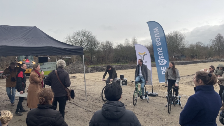 Mensen verzamelen zich onder een tent bij een open terrein; twee personen fietsen op hometrainers naast vlaggen.