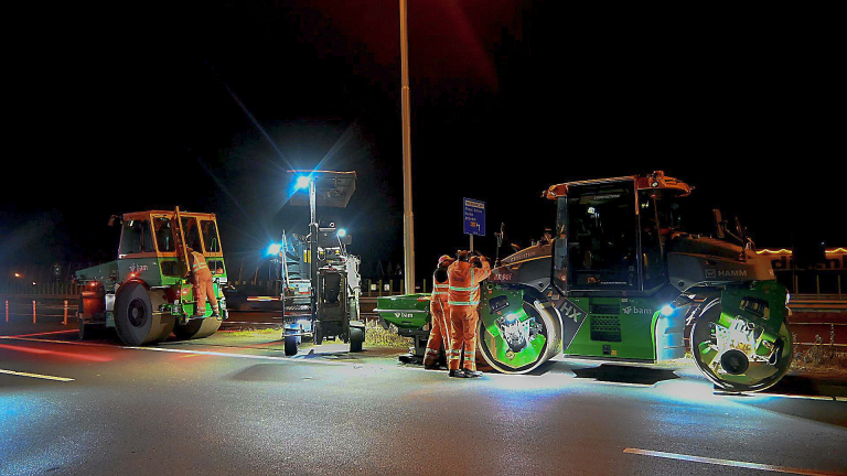 Wegenwerkers in oranje pakken werken 's nachts aan een weg met zware machines en felle lichten.