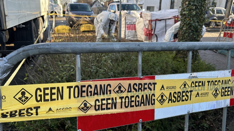 Waarschuwingslint met "Geen Toegang Asbest" markeert een afgesloten gebied; personen in beschermende kleding werken daar.