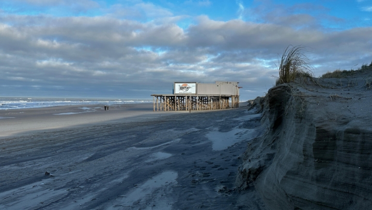 Strand met houten gebouw op palen, wolkenlucht en duinen aan de rechterkant.