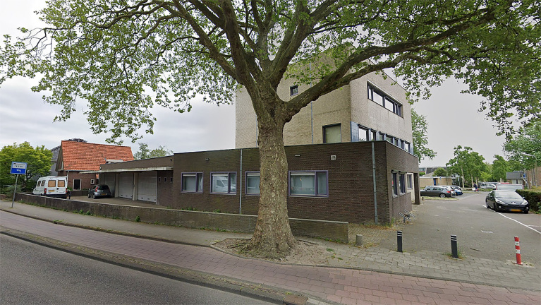 Modern gebouw met bakstenen gevel, een grote boom aan de voorzijde en een parkeerplaats met meerdere auto's.