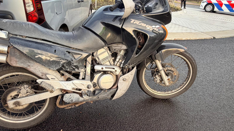 Zwarte Honda Transalp motorfiets met beschadigingen op de zijkant, geparkeerd op asfalt met een politieauto op de achtergrond.