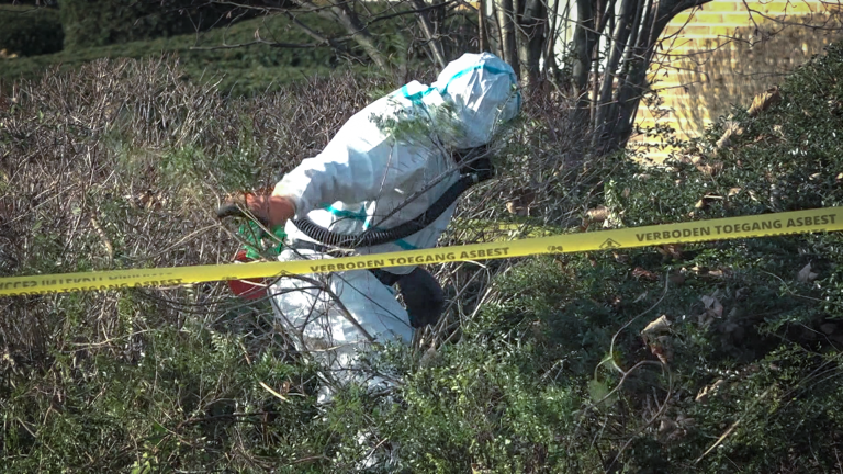 Persoon in een beschermend pak onderzoekt een gebied omgeven door lint met de tekst "Verboden toegang asbest".