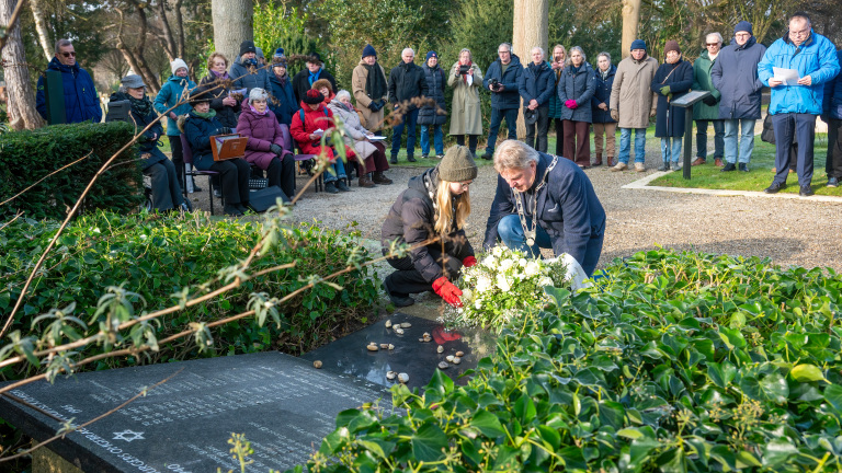 Mensen leggen bloemen op een herdenkingssteen, omringd door toeschouwers in een bosrijke omgeving.