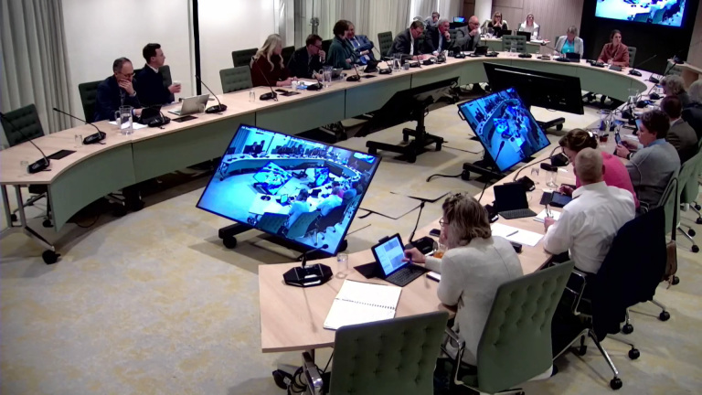 Een vergaderruimte met mensen die rond een U-vormige tafel zitten, gebruikmakend van laptops en microfoons, en schermen die de bijeenkomst weergeven.