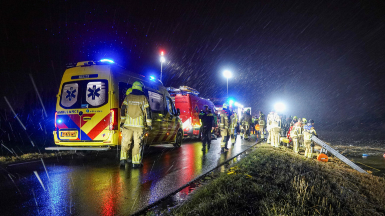 Hulpverleners werken 's nachts in de regen bij een ongeval langs de weg met een ambulance en brandweervoertuigen.