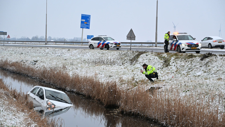 Auto deels ondergedompeld in een sloot naast een besneeuwde weg, met politiewagens en agenten die de situatie onderzoeken.